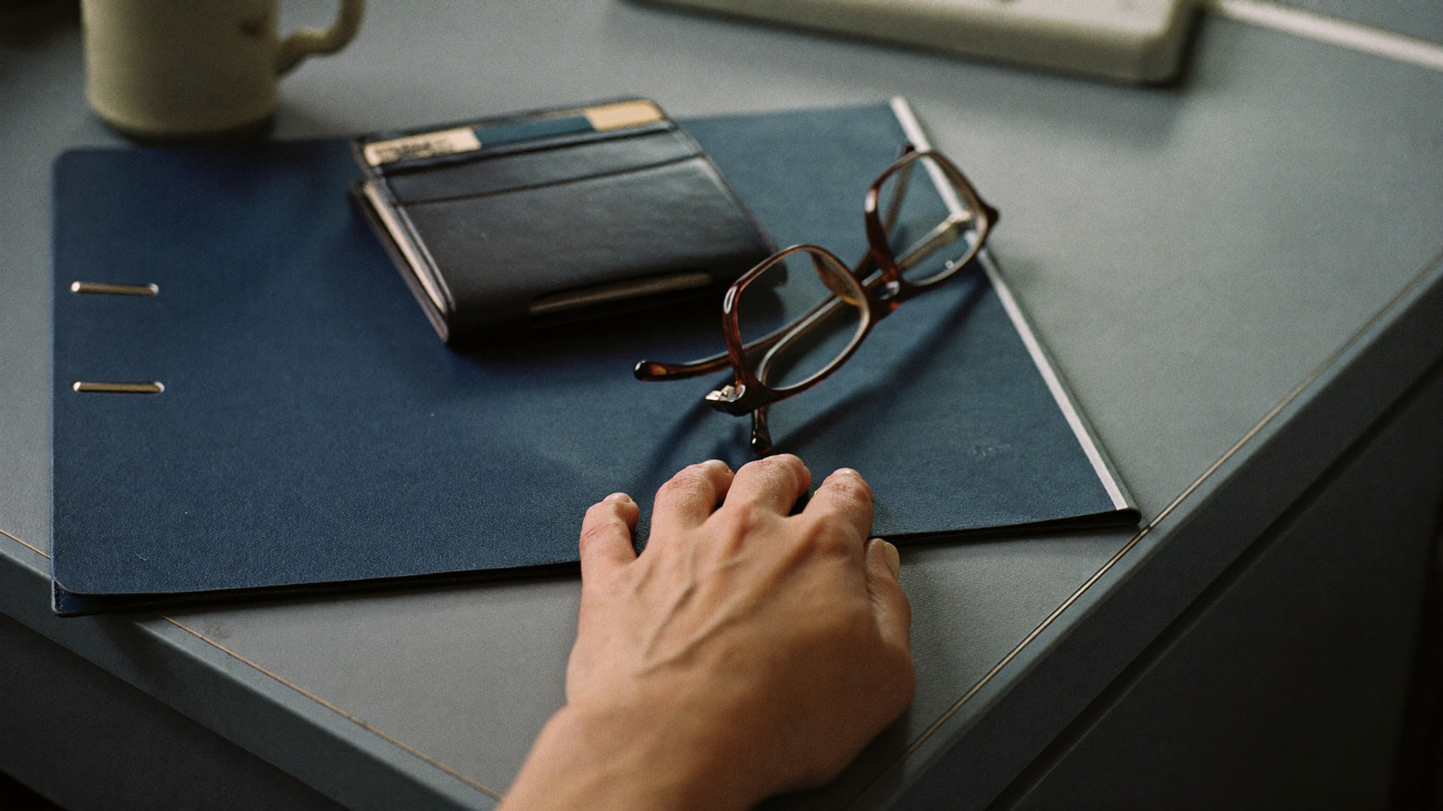 Hand liegt auf einem geschlossenen Aktenordner, darauf eine zugeklappte Dokumentenmappe und eine Lesebrille