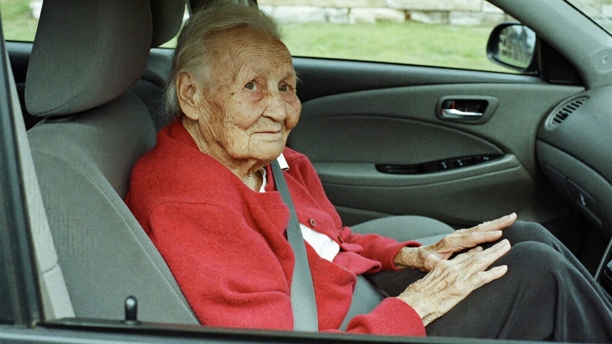 Ältere Frau sitzt angeschnallt auf dem Beifahrersitz eines Autos und wartet ruhig auf die Fahrt zum Arzt