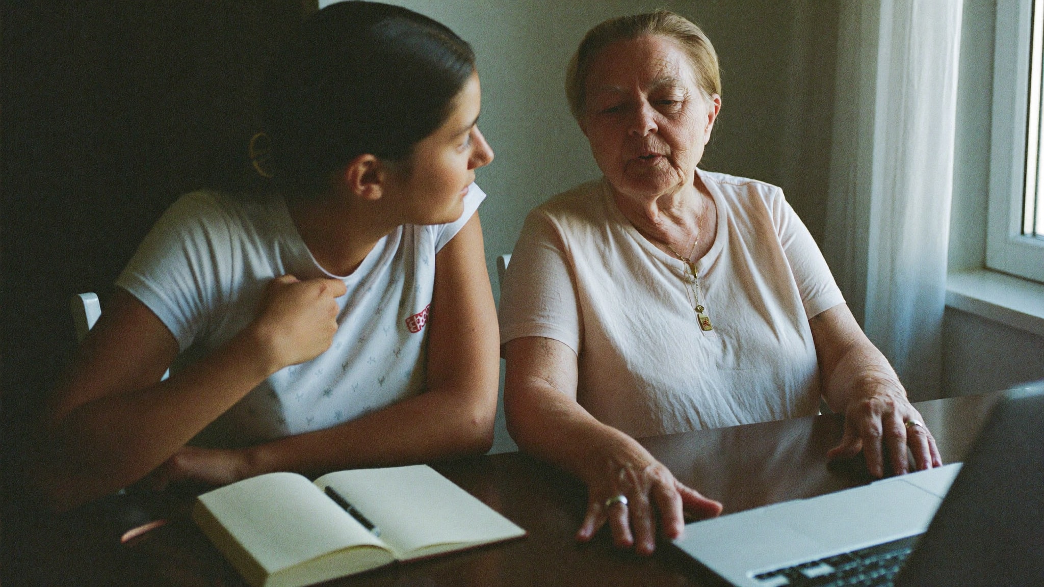 Erwachsene Tochter und ihre ältere Mutter sitzen am Esstisch vor einem Laptop und besprechen etwas