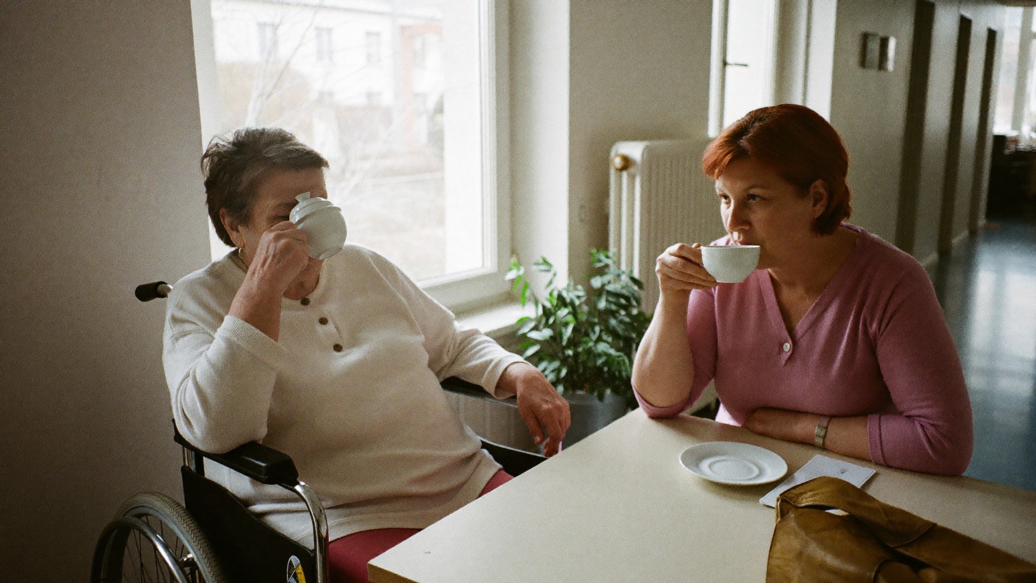 Ältere Frau im Rollstuhl und ihre Tochter sitzen gemeinsam bei einer Tasse Tee im Aufenthaltsraum einer Rehaklinik