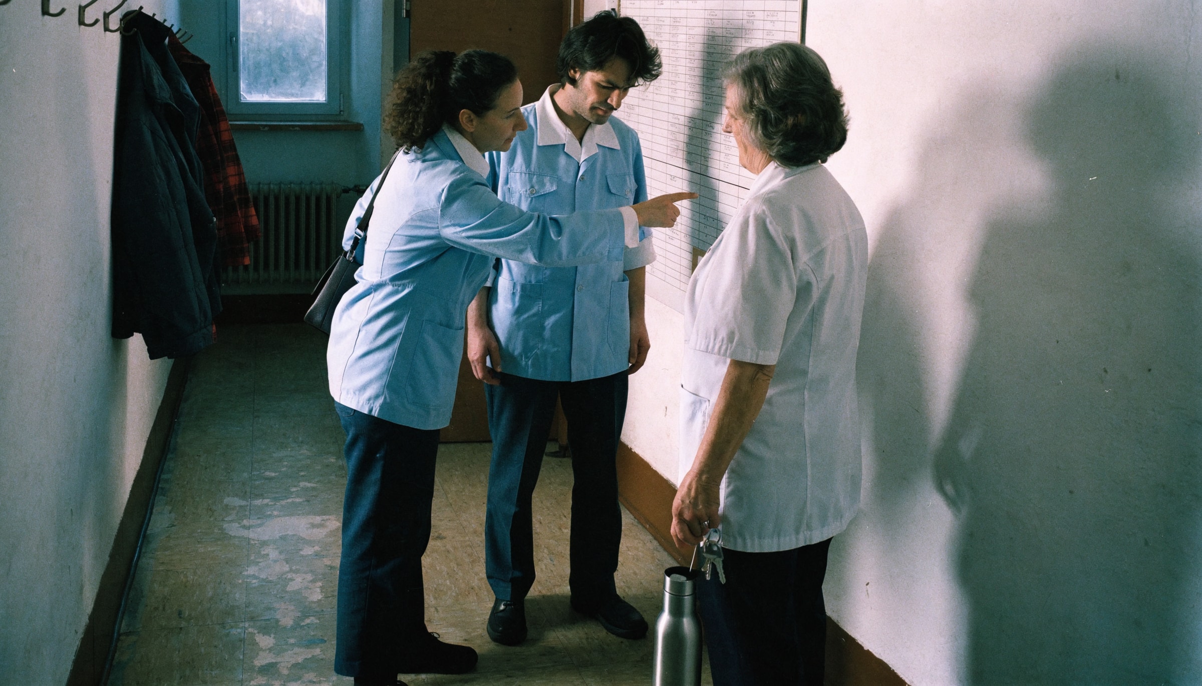 Drei Pflegekräfte stehen morgens vor dem Dienstplan an der Wand eines ambulanten Pflegedienstes und besprechen die Touren
