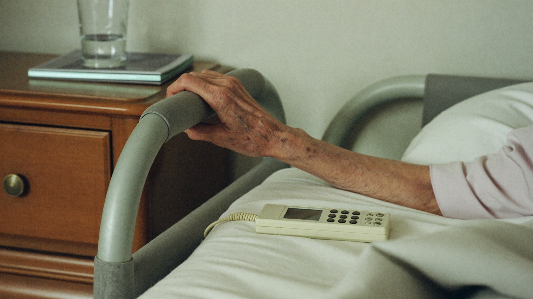 Hand einer älteren Person auf dem Seitengitter eines Pflegebetts, daneben liegt die Handbedienung auf der Decke