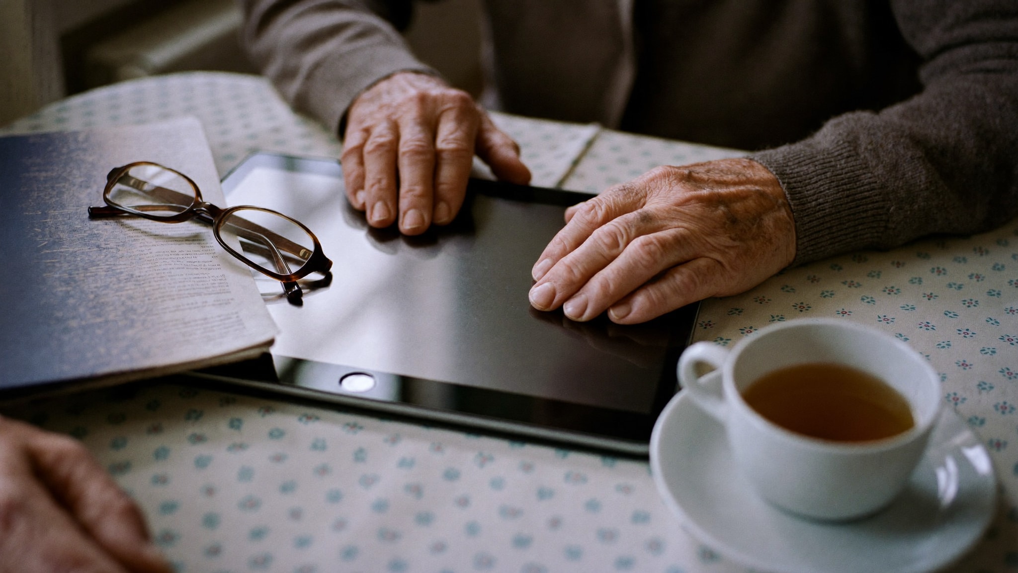 Hände einer älteren Person ruhen auf einem Tablet am Küchentisch, daneben eine Tasse Tee und eine Lesebrille