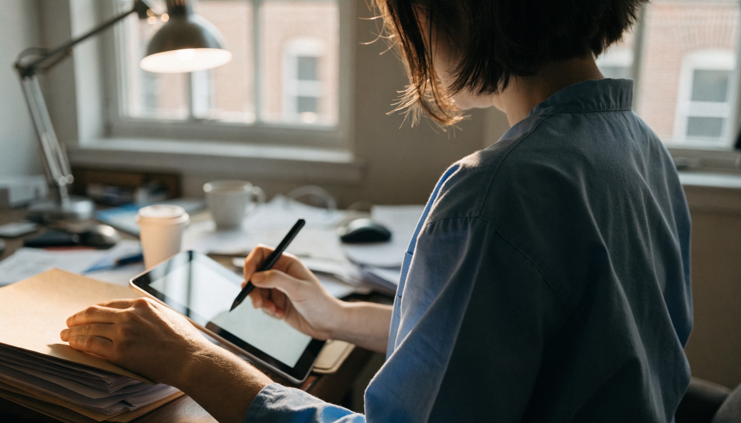 Pflegefachperson sitzt am Schreibtisch und korrigiert mit einem Eingabestift einen Textentwurf auf einem Tablet, daneben eine Papiermappe
